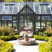 Campania Esplanade Two Tier Fountain in Cast Stone FT-78 flowing in a garden with a large glass greenhouse