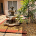 Fiore Stone 3 Tier Lion Fountain in cast stone with basin, flowing in a patio courtyard on pebbles beside a palm plant