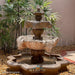 Fiore Stone 3 Tier Lion Fountain in cast stone flowing in a courtyard setting with a river rock base and green plants