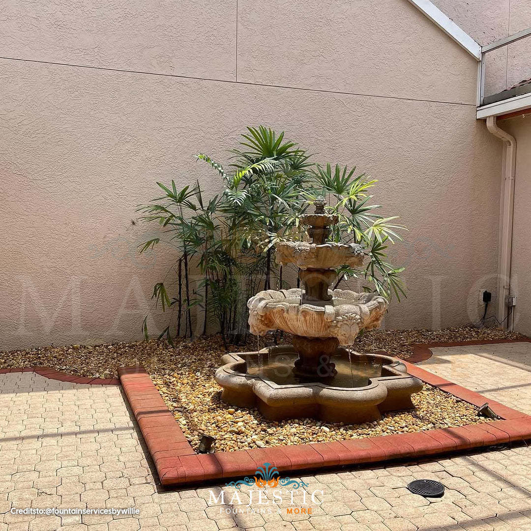 Fiore Stone 3 Tier Lion Fountain in cast stone with basin set in a courtyard pebble bed bordered by brick and pavers