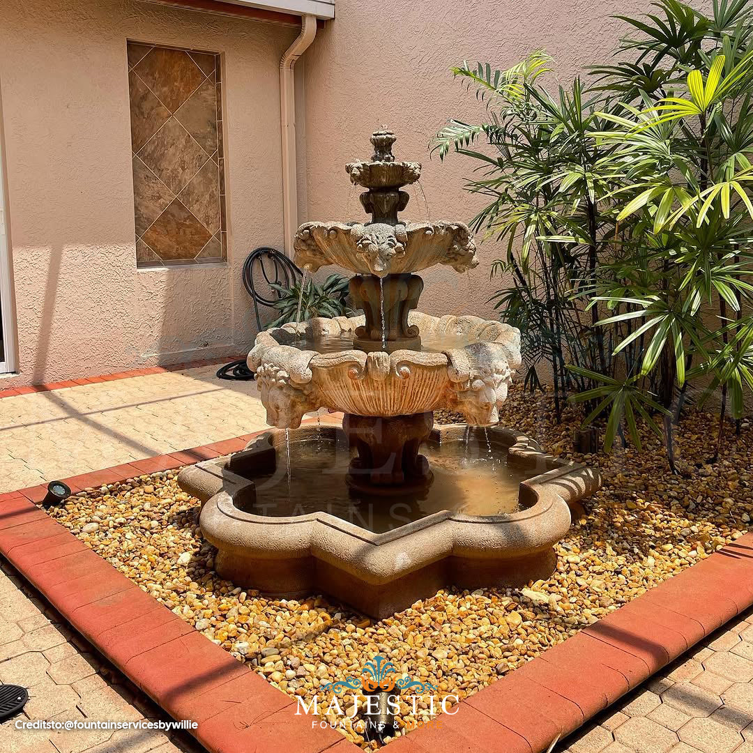 Fiore Stone 3 Tier Lion Fountain in cast stone with basin, installed in a patio rock bed beside tropical palm plants