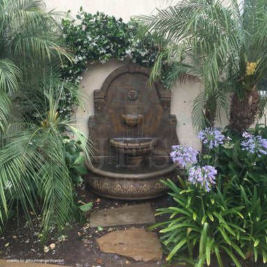 Fiore Stone Bavarian Wall Fountain in cast stone set in a garden framed by a flowering vine, palms and purple blooms