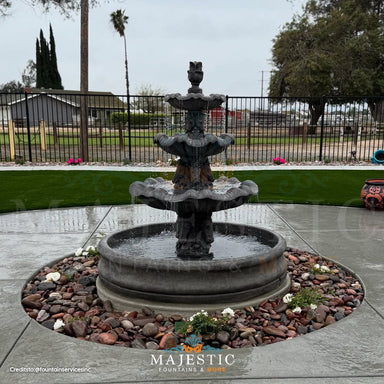 Fiore Stone Italian 3 Tier Fountain with 55 inch Basin IT-B3 in cast stone on a concrete patio surrounded by rocks